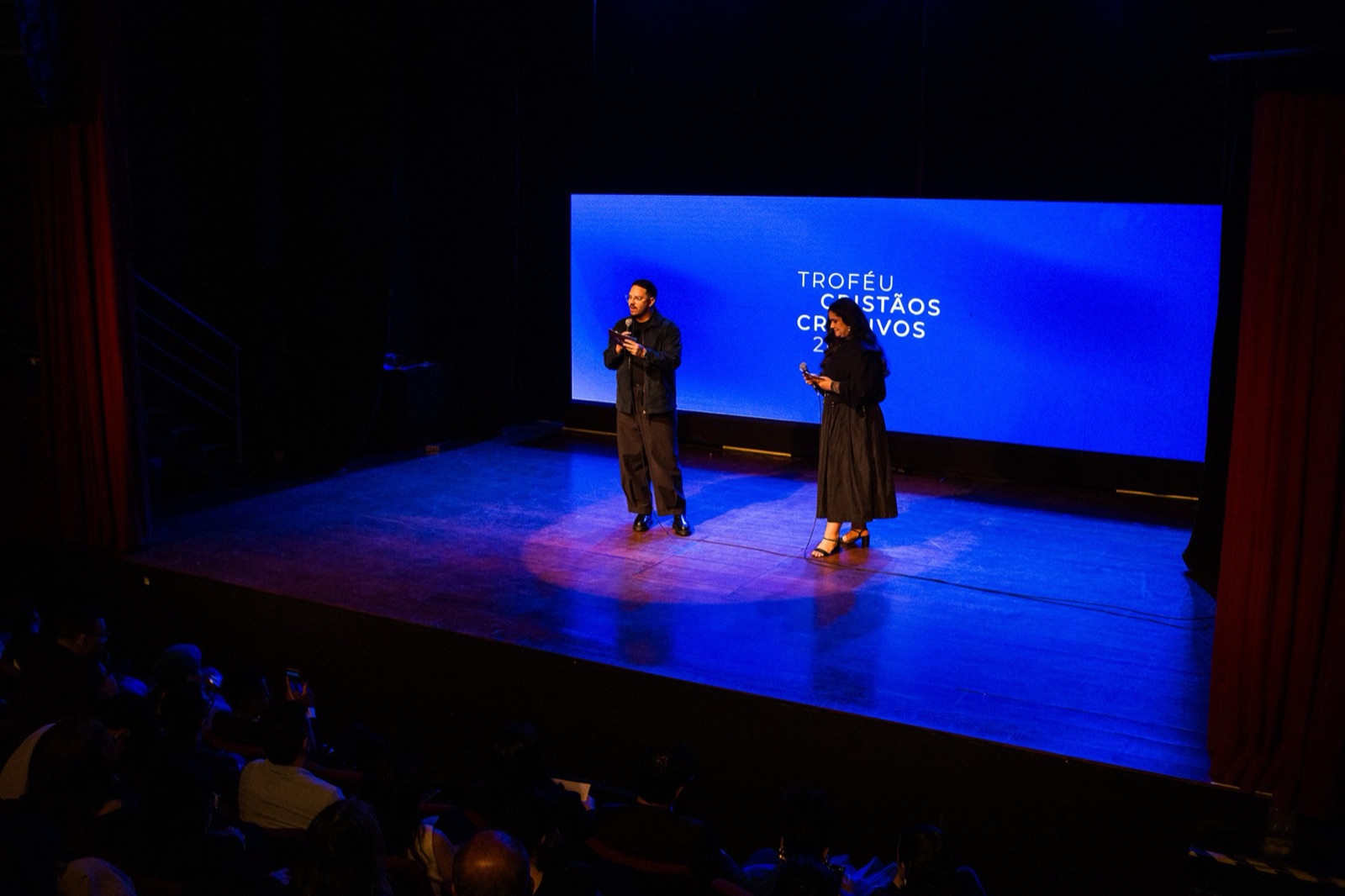 Apresentadores no palco do Teatro Viradalata, em São Paulo, diante do telão azul do Troféu Cristãos Criativos 2026, com a plateia em primeiro plano